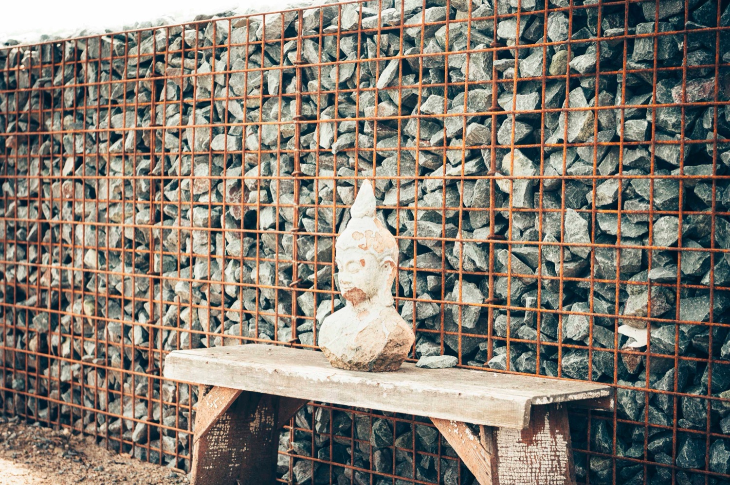 Stone bust on a reclaimed-wood bench against a gabion wall